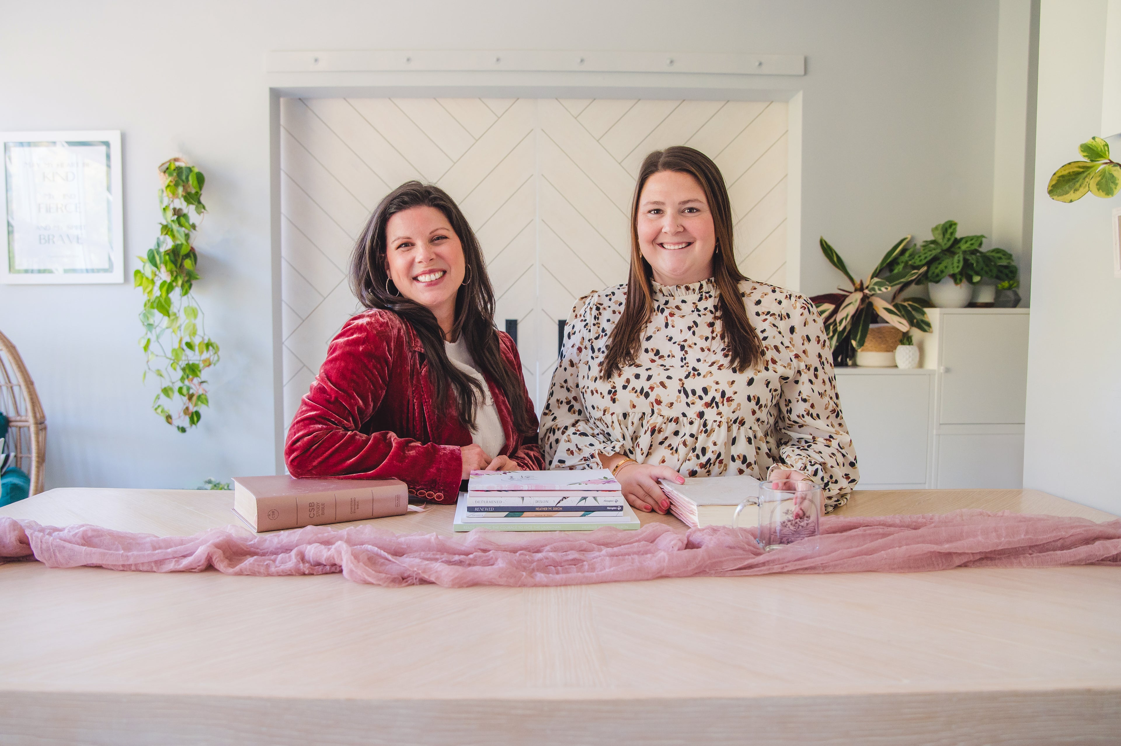 Two women sitting at a desk with a pink blanket and laptop in a home office setting.
