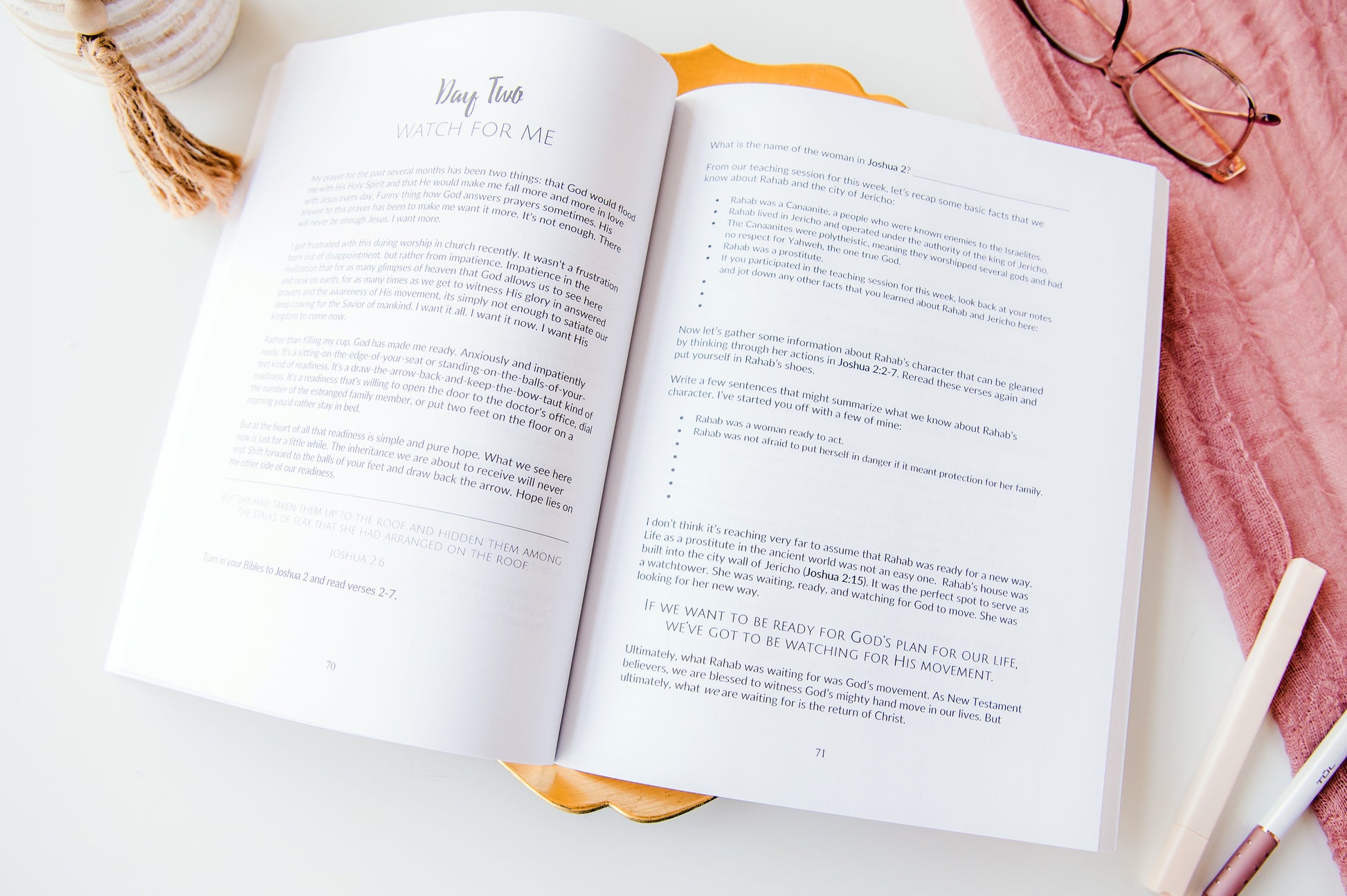 Open book on a white surface with a pink knitted blanket and glasses in the background